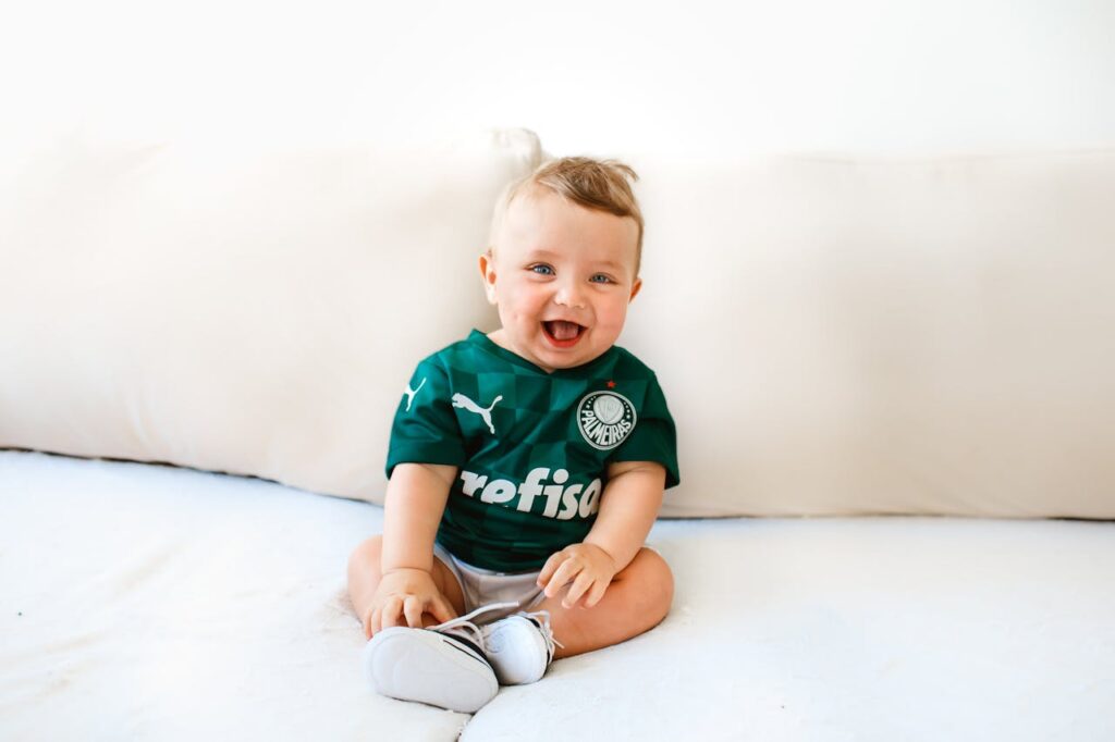 pexels-photo-9189202 Cute baby smiling while sitting on a sofa wearing a green sports jersey indoors.